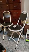 Two folding stools with white metal frames, black padded round seats and backrests, set against wooden furniture and carpeted floor.