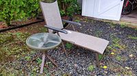 Chaise lounge with beige mesh fabric and side table with round glass top placed outdoors on stone ground in front of white shed.
