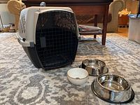 Petmate pet crate positioned on carpet next to three pet bowls (two stainless steel and one ceramic)