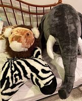 Photo showing zebra, lion, and elephant oversized plush safari animal head wall hangings arranged on a bed, showing plush fabric texture and detail.