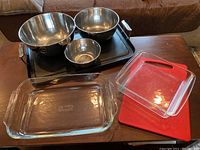 Photo showing all items in the lot arranged on a table: two Pyrex glass baking dishes front center and right, three stainless steel Crate & Barrel mixing bowls stacked on the black All-Clad griddle pan, IKEA red cutting board under a Pyrex dish.