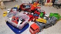 Overview of collection showing a red Mini Radio Flyer wagon, toy cars, a car hauler truck with a yellow bus on top, and a green garbage truck scattered on carpeted floor.