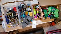 Wide view of three boxes containing assorted toys including action figures, wind-up toys, and toy weapons.