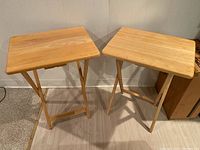 Two wooden folding tray tables side by side on floor against wall, showing rectangular tops and X-frame folding legs.