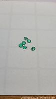 Six small round cut natural Columbian emerald gemstones on a white grid background, showing size and clarity.