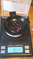 Multiple small deep pink to purple round faceted rhodolite garnet gemstones displayed in a black scoop on a precision scale showing 3.025 carats total weight.