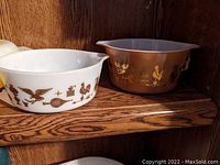 Two Pyrex casseroles side-by-side on wooden shelf, one white with brown print and one brown with gold print