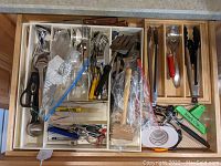 Photo showing the open kitchen drawer with stainless steel flatware forks, spoons, and knives arranged in organizer trays. Various kitchen tools including scissors, peelers, spatulas, tongs, meat tenderizer and other utensils also visible.