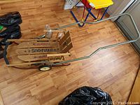 Full side view of the Gladding Sno-Flake small child push sled showing the wooden runners, metal push handle with grip, and small black wheels underneath.