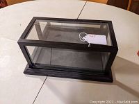 Rectangular glass display case sitting on a white round table, showing its side view and lid fixed on top.