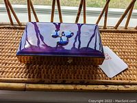 Rectangular decorative box on a woven rattan surface, showing the purple enamel lid with blue accents.