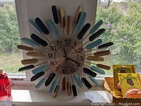 Full view of mid-century style sunburst wall clock in front of window, showing colorful accents and circular metallic clock face.