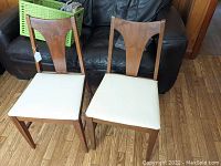 Pair of wooden mid-century style side chairs with T-shaped backrests and white upholstered seats, presented on wooden floor in front of black leather couch.