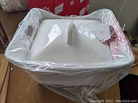 Front view of the square white ceramic casserole dish with lid, partially wrapped in plastic showing the overall shape and condition.