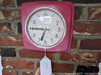 Front view of Skytimer retro style pink wall clock with clear plastic face and visible clock hands, mounted on brick wall.