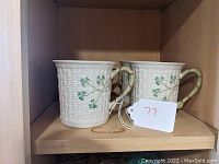 Two Belleek porcelain coffee mugs with textured basketweave pattern and green shamrock decoration displayed on wooden shelf.