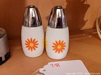 Front view of the two white glass salt and pepper shakers with orange and yellow Daisy flower pattern and shiny metal tops.