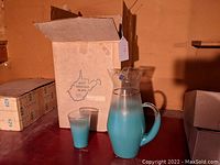 Photo of the glass pitcher and a single glass with the original box in the background showing West Virginia Glass logo and state outline