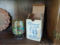 Full view of the glass candleholder next to its original box, showing the colorful embedded glass pieces and the box branding.