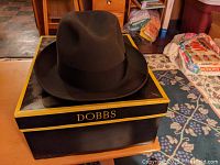 Black Dobbs fedora hat sitting on original Dobbs box showing front side with hat's pinched crown and grosgrain ribbon band.