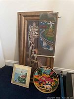 Five pieces of art arranged on floor: framed giraffe print, tapestry with Christ the Redeemer scene, framed lighthouse print, decorative plate, and wood art