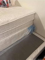 Close-up side view of mattress and box spring stack with plastic protective cover on bottom mattress.