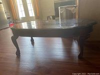 Full view of coffee table showing rectangular top with glass insert and carved wooden legs, highlighting scratches on the surface.