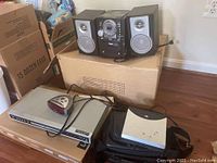 Photo showing Naxa mini stereo system with two detachable speakers, alarm clock, portable DVD player and Magnavox DVD player on top of cardboard boxes in a room.