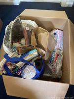 Cardboard box filled with aluminum pans, paper cups, aluminum foil, straws, and brown paper bags in reusable blue bags.