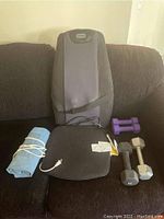 Photo showing heating pad, hand weights, and Homedics massage cushion on a couch. The heating pad is rolled and blue, with an electrical cord. The massage cushion is black and gray with attached remote. Three hand weights are visible: two gray 10 lb dumbbells and one pair of small purple dumbbells.