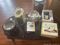 Photo showing all six appliances on a table: black Rival crockpot with glass lid, white Oyster rice cooker, Black & Decker coffee maker box, Style Elements mini sewing machine box with accessories, and Hamilton Beach juicer with GE burner visible