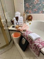 Photo showing white folded towels stacked on tiled ledge beside white bathtub, pink and white patterned towel draped, black bathroom scale on floor, small wastebasket inside bathtub corner, and metal candle holders.