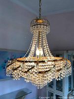 Full view of crystal beaded chandelier hanging from chain with gold metal frame and multiple light bulbs lit.