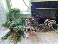 Wide view showing 12 faux flower and greenery arrangements with various pots and vases, set on a tiled floor and near a fireplace.