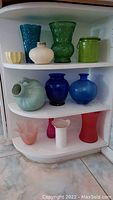 Shelf with 12 colorful vases made of various materials and shapes including tall blue pressed glass, green textured glass, and ceramic pieces.