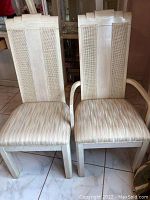 Two whitewashed wooden chairs with woven cane backs and beige patterned cushion seats shown side by side on tile floor.