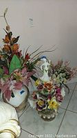Porcelain vase with colorful flower embellishments and woman figurine, alongside large white vase with faux flowers