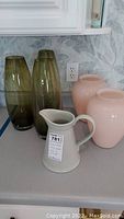 Photo showing three tall green cylindrical glass vases, two pink ginger jar-shaped ceramic vases, and a green ceramic pitcher with a handle.