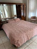 Full view of the bed made with pink bedding showing the headboard with mirror and light units, flanked by windows and chairs.