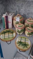 Photo showing four ceramic canisters with palm tree designs and lids, next to two matching ceramic wall plaques with palm tree and white fence motifs, and several cookbooks standing upright.