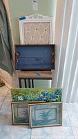 Lot of assorted home decor items including framed padded cork board, wooden side table, blue wooden serving tray, and three framed paintings arranged on a floor with curtain backdrop.