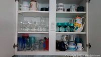 Open kitchen cabinet shelves showing various glass and ceramic drinkware including a ceramic pitcher with fruit decoration, clear and green wine glasses, colored drinking glasses, baking dishes, and mugs.