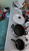 Photo showing five metal pots, one large lid, and metal baking pans on a table. Items appear used with scratches.