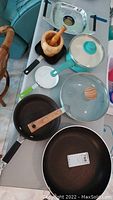 Top view showing five frying pans of various sizes and materials, one green pot with glass lid, and wooden mortar and pestle arranged on table.