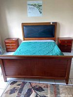 Full view of queen bed frame with headboard featuring black padded panel and wooden footboard. Teal bedding present, mattress not included.