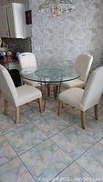 Round glass dining table with four cream upholstered chairs surrounding it, tiled floor, light floral wallpaper background.