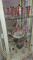 Glass cabinet shelf showing 6 pink and gold-tone vintage champagne flutes, porcelain flower-basket, and assorted decorative bottles below.