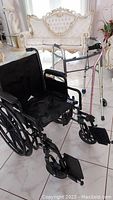 Photo showing the black Drive wheelchair next to the silver walker and canes on a tiled floor in a living room setting.