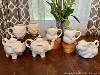 Wide shot showing all 7 ceramic figural pieces in white: 2 owl mugs, 2 fox mugs, 2 elephant teapots, 1 rabbit teapot.