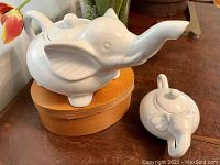 Elephant-shaped white ceramic teapot and creamer displayed on wooden surface with tulips in background.
